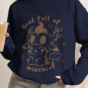 Peut inclure: Un sweat-shirt bleu marine à manches longues avec une impression graphique marron clair représentant un personnage de dessin animé portant une coiffe à cornes. Le texte "Head full of mischief" est imprimé en arc autour du personnage.