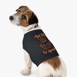 Peut inclure: Un t-shirt noir pour animal de compagnie avec un motif orange représentant une créature ressemblant à un hibou à l'intérieur d'une citrouille. Le texte "Too cute to spook" est imprimé en orange avec de petites étoiles décoratives.