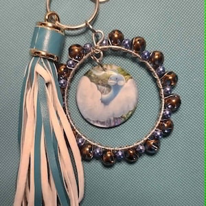 Peut inclure: Un porte-clés composé d'un anneau en métal argenté entouré de fil métallique avec des perles marron foncé et de petites perles bleues. Un médaillon circulaire illustrant une créature bleue est suspendu. Un pompon en similicuir bleu canard et blanc.
