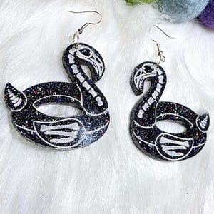May include: A pair of black glitter flamingo earrings with white skeleton detailing. The earrings have a hook closure. The earrings are set against a white, fluffy background. Colourful felt balls are scattered around.