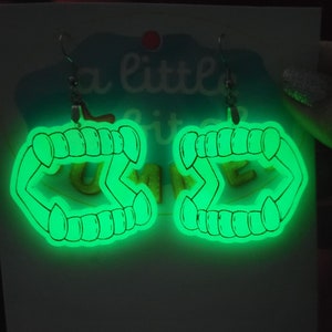 May include: A pair of glow-in-the-dark earrings shaped like vampire fangs. The earrings are green and have a black outline. The earrings are hanging from silver hooks.