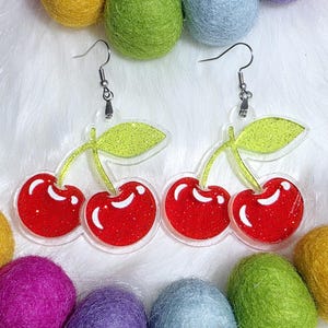 Puede incluir: Pendientes con forma de cereza con cerezas rojas y hojas verdes, hechos de acrílico transparente con ganchos plateados. Bolas de fieltro de colores en tonos morado, amarillo, verde y azul rodean los pendientes.