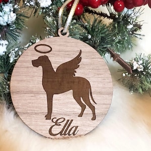 K&ouml;nnte beinhalten: Ein h&ouml;lzerner Weihnachtsbaumschmuck mit einer Silhouette eines Hundes mit Engelsfl&uuml;geln und einem Heiligenschein. Der Anh&auml;nger ist mit dem Namen "Ella" personalisiert.