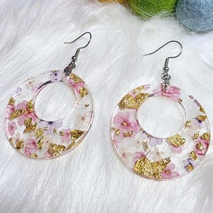 May include: A pair of clear, circular earrings with a floral design and gold leaf accents. The earrings feature a silver hook and are filled with pink and white flowers. The earrings are displayed on a white, fluffy surface.