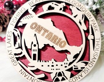 May include: A circular wooden ornament featuring a cutout map of Ontario with the text ONTARIO. The border includes engraved text: BEAVER TAIL, ALGONQUIN PARK, THOUSAND ISLANDS, BLUE MOUNTAIN, GREAT LAKES, FISHING. It has a red backing and a top hole.