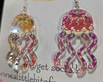 May include: A pair of dangling jellyfish earrings made of translucent acrylic. One earring features a yellow and orange gradient, while the other displays a pink and purple gradient. Both have white floral and dot patterns and silver hooks.