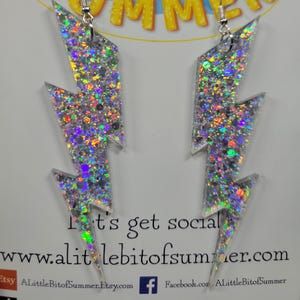 May include: A pair of silver glitter lightning bolt earrings. The earrings are made of a clear material filled with iridescent glitter. The earrings hang from silver hooks. The background has the text "a little bit of SUMMER" and "Let's get social".