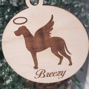 K&ouml;nnte beinhalten: Ein Holz-Anh&auml;nger mit einer Hundesilhouette mit Engelsfl&uuml;geln und einem Heiligenschein. Der Hund ist braun und der Anh&auml;nger ist hellbraun. Der Anh&auml;nger tr&auml;gt den Namen "Breezy" eingraviert.