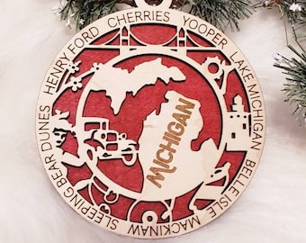 Puede incluir: Un adorno redondo de madera con fondo rojo y un recorte de madera clara del estado de Michigan. El diseño incluye un coche, un puente y el texto Henry Ford, Cherries, Yooper, Lake Michigan, Belle Isle, Mackinaw, Sleeping Bear Dunes.