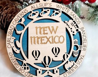 May include: A circular wooden ornament featuring a teal background and laser-cut details. The centre displays the text NEW MEXICO above three hot air balloons. The outer border lists landmarks including CARLSBAD CAVERNS, ALLSUP'S, WHITE SANDS, BANDELIER, TAOS PUEBLO, KOKOPELLI, and RED OR GREEN?