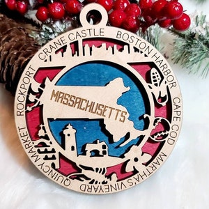 May include: A wooden Christmas ornament shaped like the state of Massachusetts with the text "Massachusetts" in gold lettering. The ornament is painted in shades of red, blue, and brown, and features various landmarks and locations around the state, including Boston Harbor, Cape Cod, Martha's Vineyard, and Quincy Market. The ornament is decorated with a gold rope hanger.