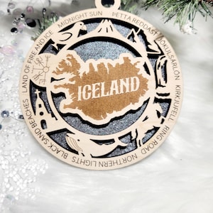 May include: A circular wooden ornament featuring a cutout map of Iceland with the text ICELAND in the center. The border includes engraved text and icons representing landmarks like a church, whale, and volcano, set against a dark glittery background.