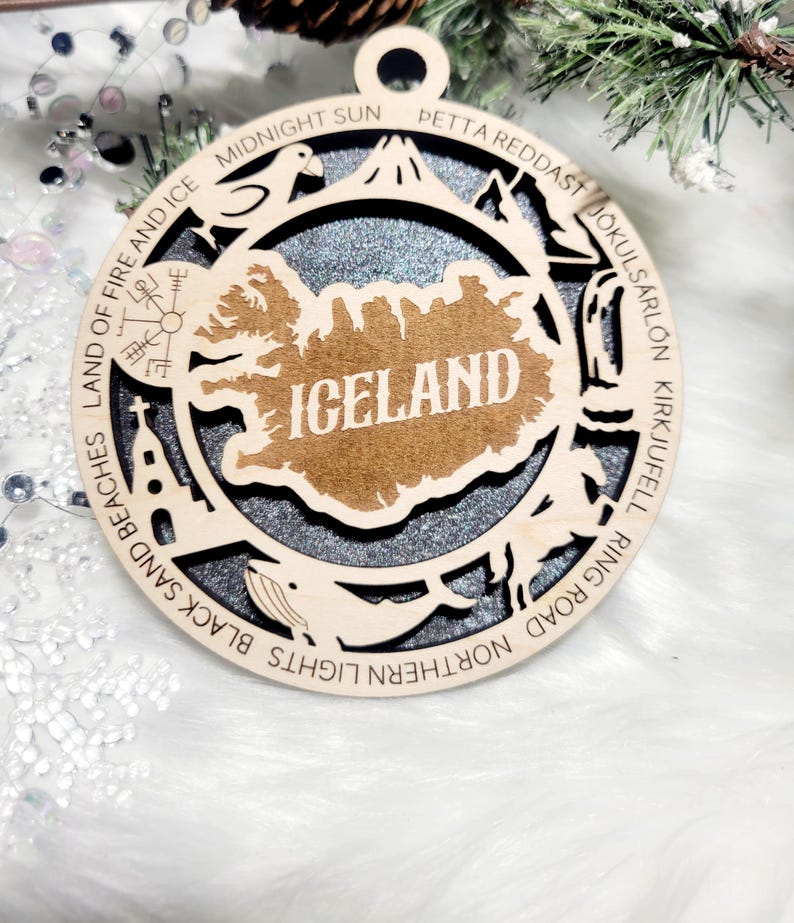 May include: A circular wooden ornament featuring a cutout map of Iceland with the text ICELAND in the center. The border includes engraved text and icons representing landmarks like a church, whale, and volcano, set against a dark glittery background.