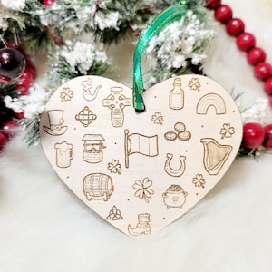 May include: A heart-shaped wooden ornament with engraved St. Patrick's Day themed icons, including a flag, shamrocks, and a pot of gold. It has a green ribbon for hanging and is displayed against a festive background.