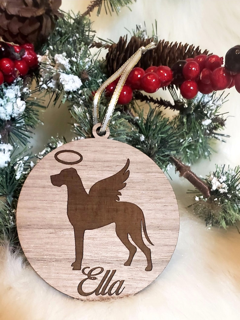 K&ouml;nnte beinhalten: Ein h&ouml;lzerner Weihnachtsbaumschmuck mit einer Silhouette eines Hundes mit Engelsfl&uuml;geln und einem Heiligenschein. Der Anh&auml;nger ist mit dem Namen "Ella" personalisiert.