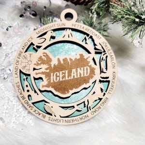 May include: A circular wooden ornament featuring a cutout map of Iceland with the text ICELAND in the center. The border includes engraved text and icons representing landmarks like Land of Fire and Ice, Midnight Sun, and J&ouml;kuls&aacute;rl&oacute;n.