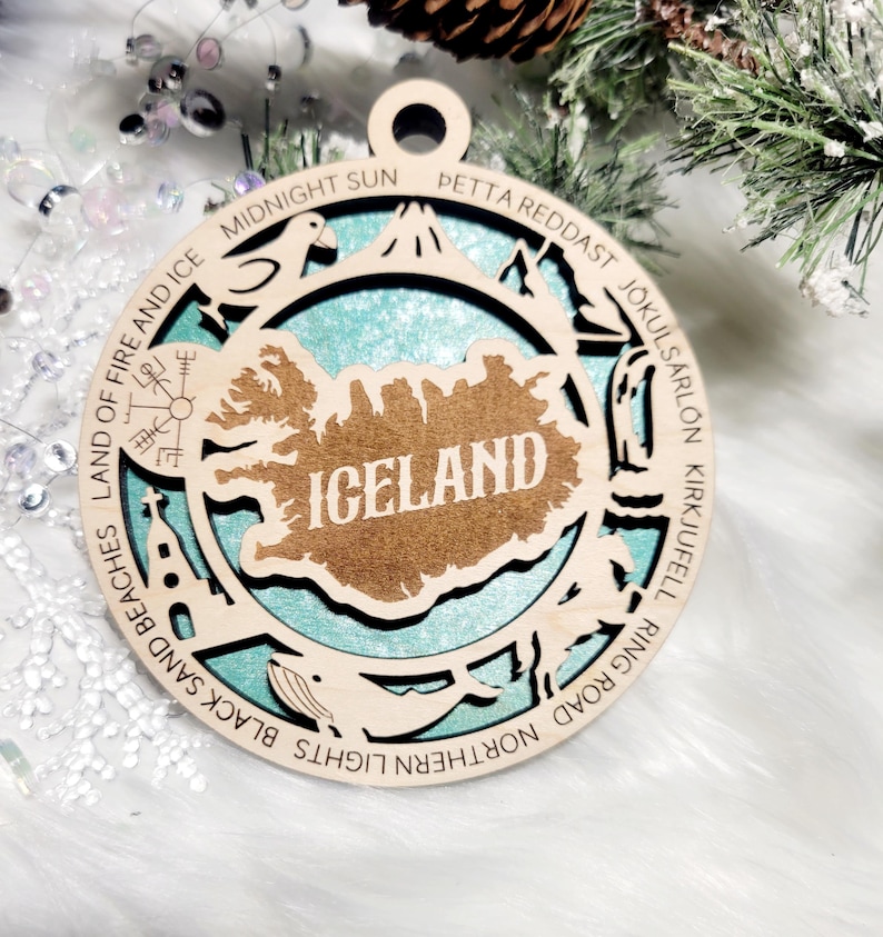 May include: A circular wooden ornament featuring a cutout map of Iceland with the text ICELAND in the center. The border includes engraved text and icons representing landmarks like Land of Fire and Ice, Midnight Sun, and J&ouml;kuls&aacute;rl&oacute;n.