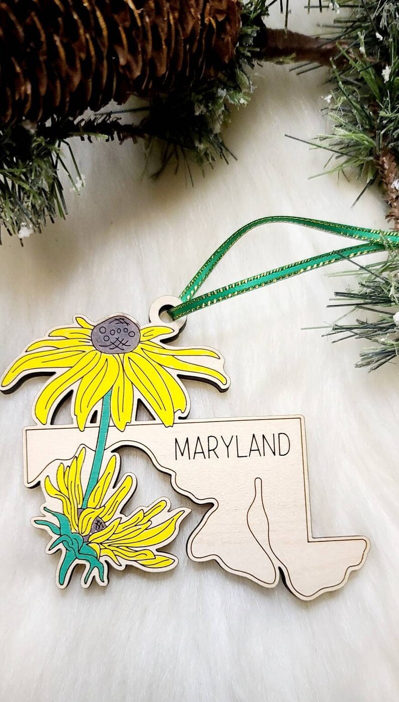May include: A wooden ornament shaped like the state of Maryland with the word MARYLAND printed in black. The ornament features two yellow black-eyed Susan flowers with green stems and a green ribbon loop for hanging from the top of the piece.
