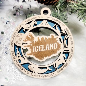 May include: A circular wooden ornament featuring a cutout map of Iceland with the text ICELAND in the center. The border includes engraved text: LAND OF FIRE AND ICE, MIDNIGHT SUN, PETTA REDDAST, J&Ouml;KULS&Aacute;RL&Oacute;N, KIRKJUFEL, RING ROAD, NORTHERN LIGHTS, BLACK SAND BEACHES.