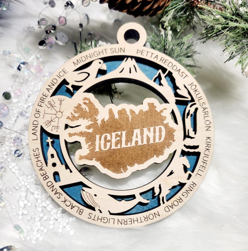 May include: A circular wooden ornament featuring a cutout map of Iceland with the text ICELAND in the center. The border includes engraved text: LAND OF FIRE AND ICE, MIDNIGHT SUN, PETTA REDDAST, J&Ouml;KULS&Aacute;RL&Oacute;N, KIRKJUFEL, RING ROAD, NORTHERN LIGHTS, BLACK SAND BEACHES.