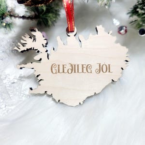 Puede incluir: Un adorno de madera con forma del contorno de Islandia con el texto "Gleðileg Jól", que en islandés significa "Feliz Navidad", colgado de una cinta roja.
