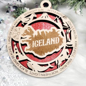 May include: A circular wooden ornament featuring a cutout map of Iceland with the text ICELAND in the center. The border includes engraved text and icons representing landmarks like a church, whale, and bird, set against a red background.