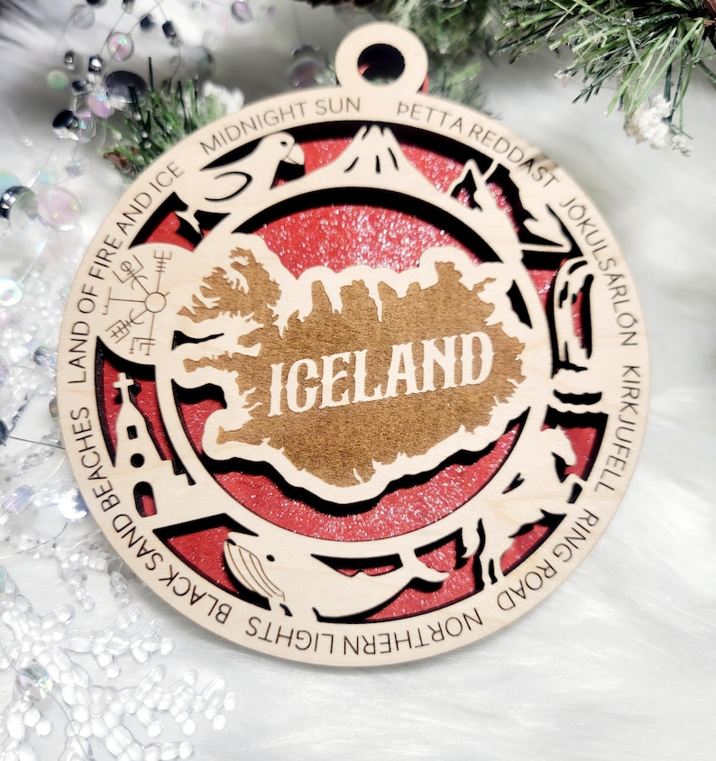 May include: A circular wooden ornament featuring a cutout map of Iceland with the text ICELAND in the center. The border includes engraved text and icons representing landmarks like a church, whale, and bird, set against a red background.