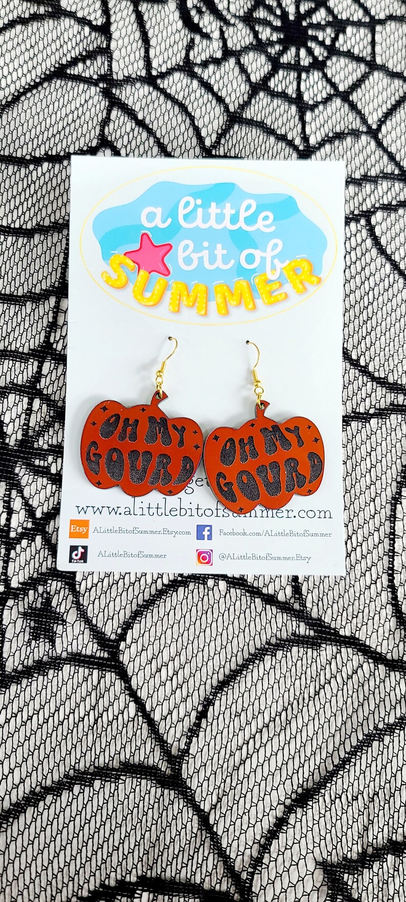 May include: A pair of pumpkin-shaped earrings with the words 'Oh My Gourd' written in black on an orange background. The earrings are hanging from gold hooks. The earrings are displayed on a white card with the words 'a little bit of summer' and a star. The card also includes a website address and social media icons.