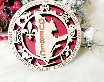 May include: A circular wooden ornament featuring a red background and a cutout map of Quebec with the text QUEBEC. The border includes the words HUDSON BAY, WINTER CARNIVAL, CHUTES MONTMORENCY, NOTRE-DAME BASILICA, MONT TREMBLANT, and POUTINE.