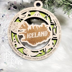May include: A circular wooden ornament featuring a cutout map of Iceland in the center with the text ICELAND. The border displays engraved text including MIDNIGHT SUN, PETTA REDDAST, J&Ouml;KULS&Aacute;RL&Oacute;N, KIRKJUFELL, NORTHERN LIGHTS, BLACK SAND BEACHES, and LAND OF FIRE AND ICE.