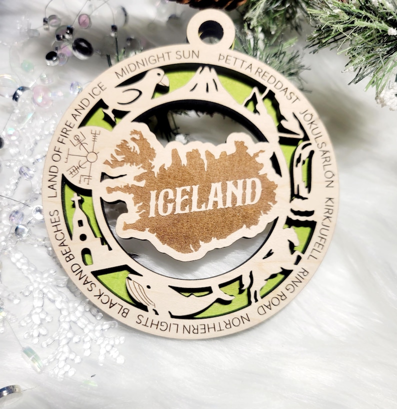 May include: A circular wooden ornament featuring a cutout map of Iceland in the center with the text ICELAND. The border displays engraved text including MIDNIGHT SUN, PETTA REDDAST, J&Ouml;KULS&Aacute;RL&Oacute;N, KIRKJUFELL, NORTHERN LIGHTS, BLACK SAND BEACHES, and LAND OF FIRE AND ICE.