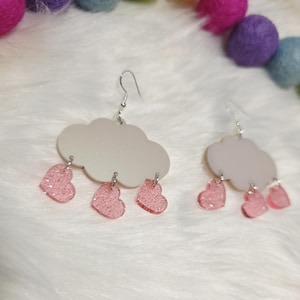May include: A pair of cloud-shaped earrings with pink heart-shaped raindrops. The earrings have silver-colored hooks and are set against a white, fluffy background. The hearts have a glittery finish.