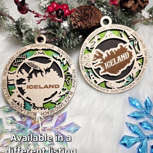 May include: Two circular wooden ornaments featuring a cutout map of Iceland with the text ICELAND in the center. The outer rings are engraved with locations like Thingvellir, Skaftafell, and Reykjavik, layered over a green and blue background.
