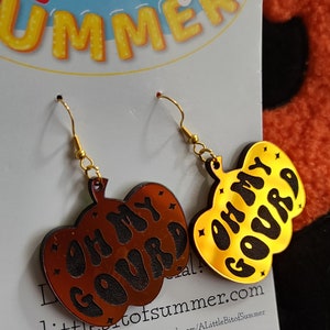 May include: A pair of pumpkin-shaped dangle earrings featuring gold-tone metal hooks. One earring is a dark brown acrylic with black text reading 'OH MY GOURD', and the other is a reflective gold acrylic with black text reading 'OH MY GOURD'.