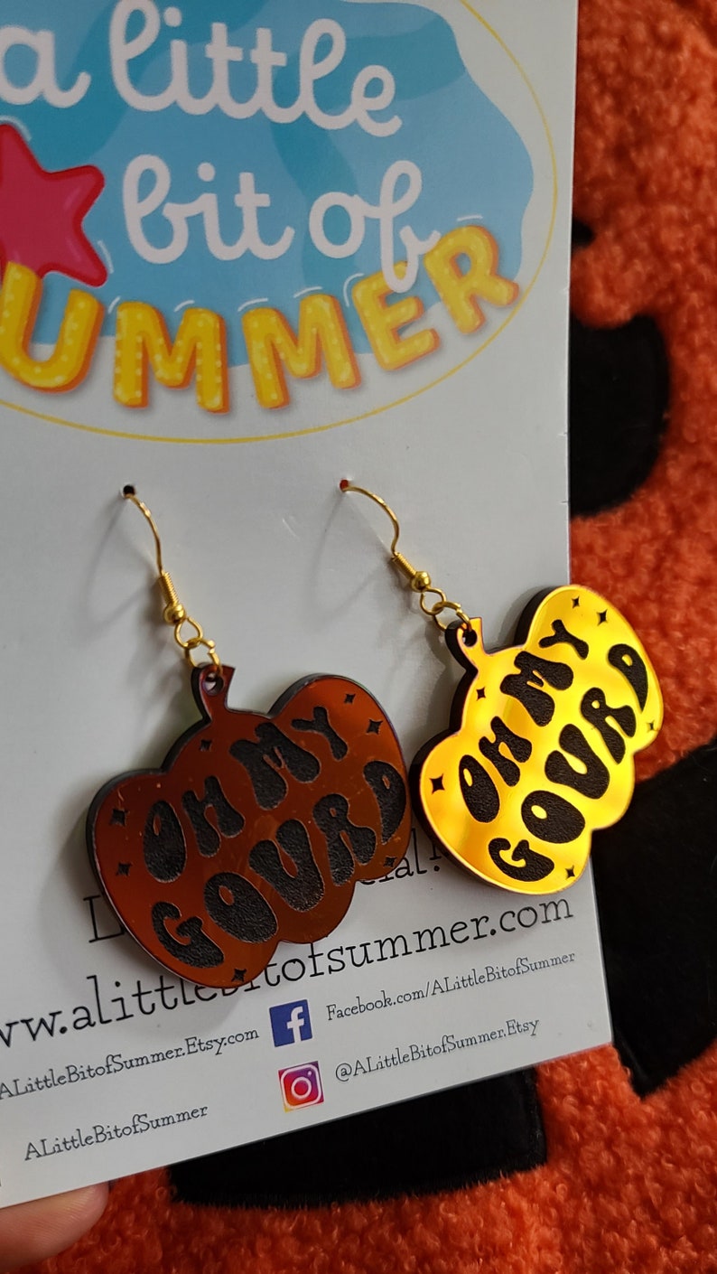 May include: A pair of pumpkin-shaped dangle earrings featuring gold-tone metal hooks. One earring is a dark brown acrylic with black text reading 'OH MY GOURD', and the other is a reflective gold acrylic with black text reading 'OH MY GOURD'.