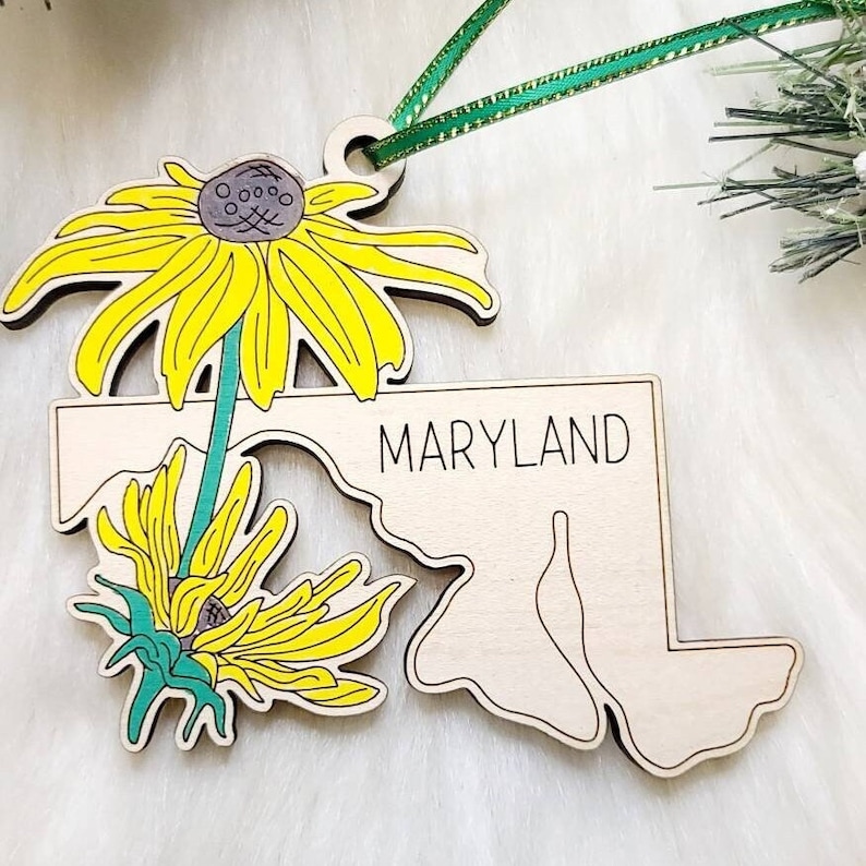 May include: A wooden ornament shaped like the state of Maryland with the word MARYLAND printed in black. The ornament features two yellow black-eyed Susan flowers with green stems and a green ribbon attached to the top for hanging purposes.