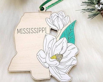 May include: A wooden ornament shaped like the state of Mississippi with the text MISSISSIPPI printed in black. The ornament features a large white magnolia flower with a yellow center and a single green leaf, attached to a green ribbon for hanging.