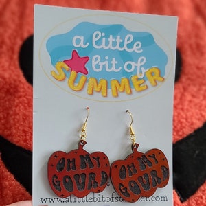 May include: A pair of pumpkin-shaped dangle earrings featuring a dark orange finish with the black text OH MY GOURD engraved on the front. The earrings hang from gold-toned metal hooks and are displayed on a white card with a blue logo.