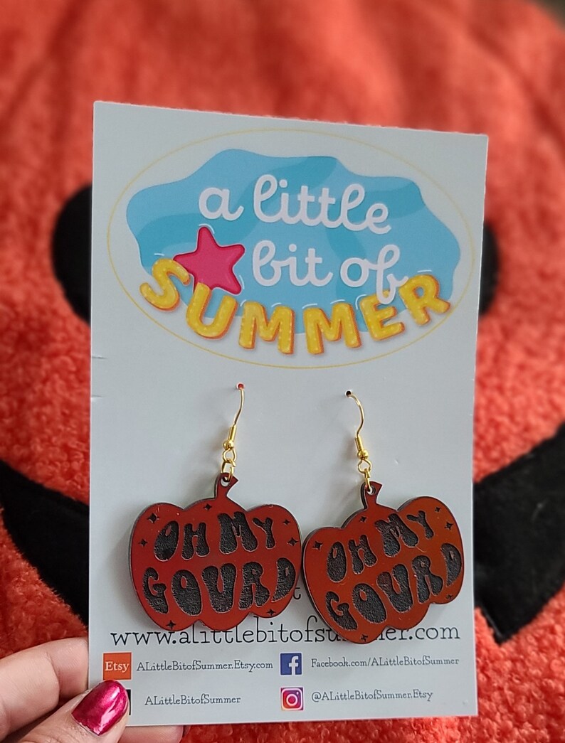 May include: A pair of pumpkin-shaped dangle earrings featuring a dark orange finish with the black text OH MY GOURD engraved on the front. The earrings hang from gold-toned metal hooks and are displayed on a white card with a blue logo.