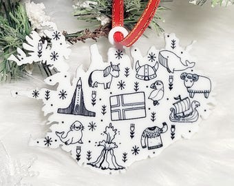May include: A white, flat, map-shaped ornament featuring black line art illustrations of Icelandic icons including a horse, a puffin, a whale, a sheep, a volcano, a church, a Viking ship, a sweater, a helmet, and the national flag, hung by a red ribbon.