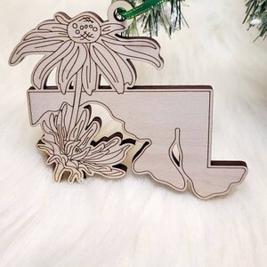 May include: A light-coloured wooden ornament shaped like the state of Maryland, featuring an engraved line art design of two coneflower blossoms. The ornament includes a small hole at the top with a thin green ribbon attached for hanging.
