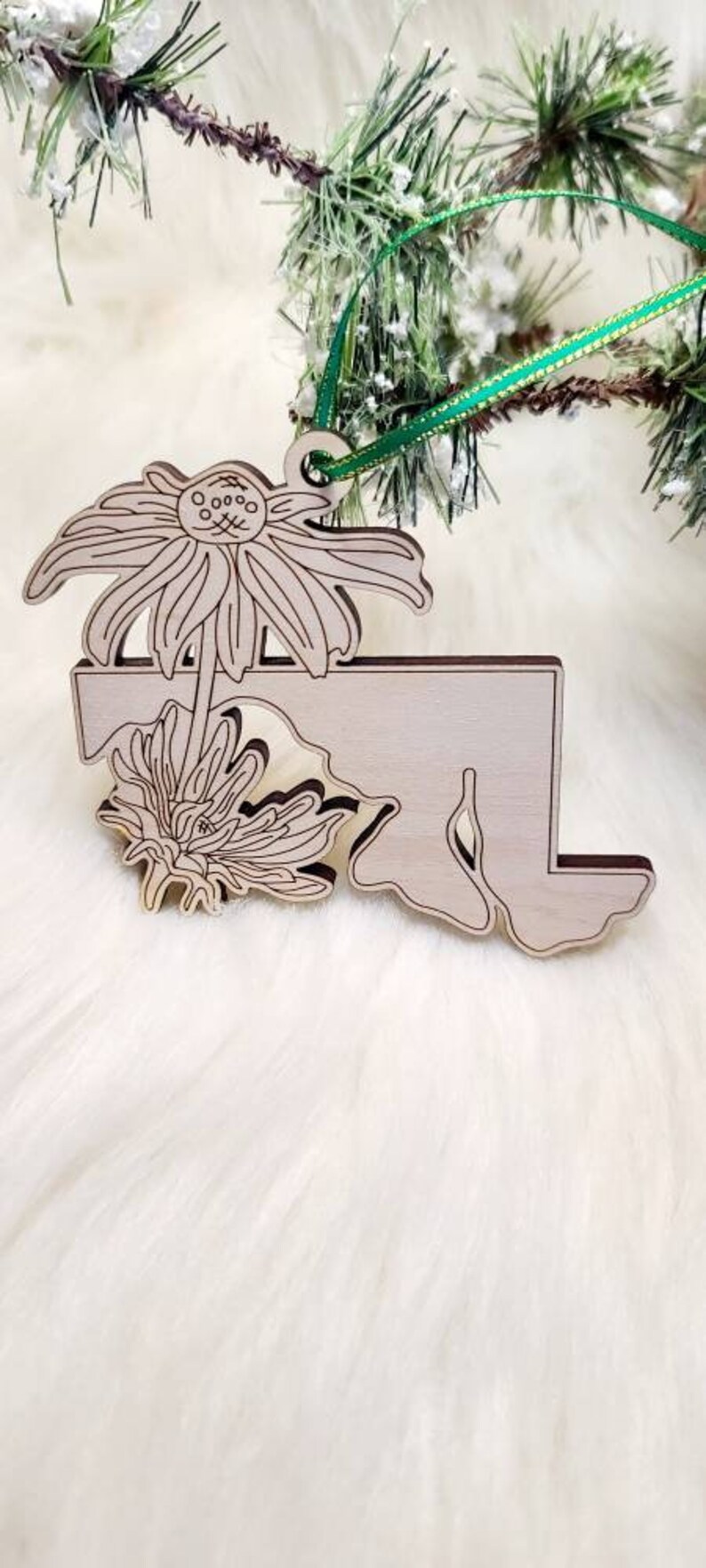 May include: A light-coloured wooden ornament shaped like the state of Maryland, featuring an engraved line art design of two coneflower blossoms. The ornament includes a small hole at the top with a thin green ribbon attached for hanging.