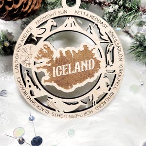 May include: A circular wooden ornament featuring a cutout map of Iceland in the center with the text ICELAND. The border includes engravings of landmarks and text: MIDNIGHT SUN, PETTA REDDAST, J&Ouml;KULS&Aacute;RL&Oacute;N, KIRKJUFELL, RING ROAD, NORTHERN LIGHTS, BLACK SAND BEACHES, LAND OF FIRE AND ICE.