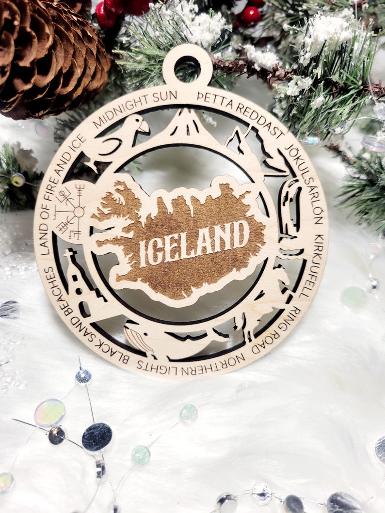 May include: A circular wooden ornament featuring a cutout map of Iceland in the center with the text ICELAND. The border includes engravings of landmarks and text: MIDNIGHT SUN, PETTA REDDAST, J&Ouml;KULS&Aacute;RL&Oacute;N, KIRKJUFELL, RING ROAD, NORTHERN LIGHTS, BLACK SAND BEACHES, LAND OF FIRE AND ICE.