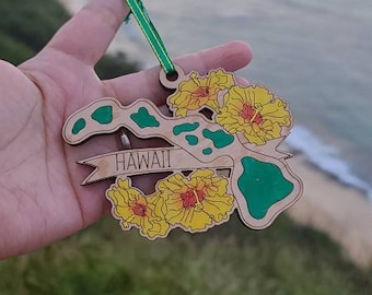 May include: A wooden ornament shaped like the Hawaiian islands, featuring green painted landmasses, yellow hibiscus flowers, and a banner with the text HAWAII. The ornament includes a green ribbon loop for hanging, showcasing detailed laser-cut edges.