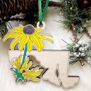 May include: A light-coloured wooden ornament shaped like the state of Maryland, featuring a vibrant yellow and green black-eyed Susan flower illustration. The ornament hangs from a thin green ribbon and has a smooth, natural wood finish.