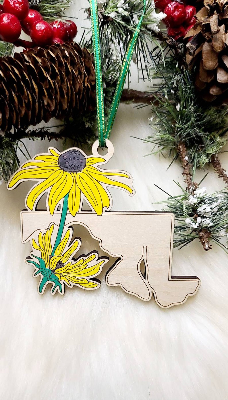 May include: A light-coloured wooden ornament shaped like the state of Maryland, featuring a vibrant yellow and green black-eyed Susan flower illustration. The ornament hangs from a thin green ribbon and has a smooth, natural wood finish.