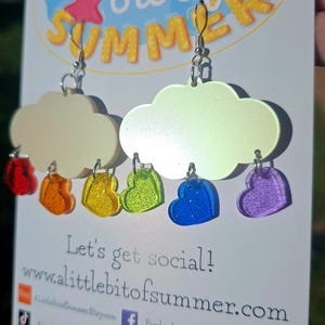 May include: Cloud-shaped earrings with dangling heart charms in red, orange, yellow, green, blue, and purple. The earrings are on a white card with the text "a little bit of SUMMER" and social media information.