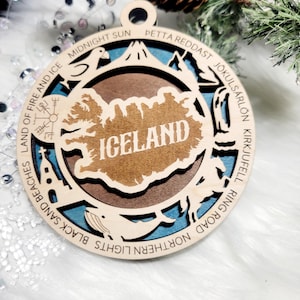May include: A circular wooden ornament featuring a layered design with a central map of Iceland. The border includes engraved text: LAND OF FIRE AND ICE, MIDNIGHT SUN, PETTA REDDAST, J&Ouml;KULS&Aacute;RL&Oacute;N, KIRKJUFELL, RING ROAD, NORTHERN LIGHTS, BLACK SAND BEACHES.