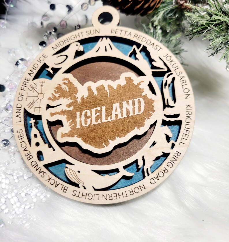 May include: A circular wooden ornament featuring a layered design with a central map of Iceland. The border includes engraved text: LAND OF FIRE AND ICE, MIDNIGHT SUN, PETTA REDDAST, J&Ouml;KULS&Aacute;RL&Oacute;N, KIRKJUFELL, RING ROAD, NORTHERN LIGHTS, BLACK SAND BEACHES.